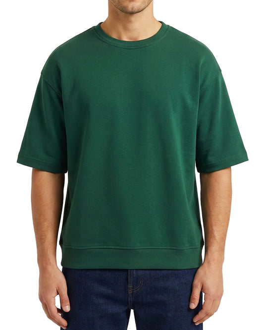 A man wearing an oversized dark green crewneck sweatshirt and dark blue jeans, shown from the neck down against a light off-white background.