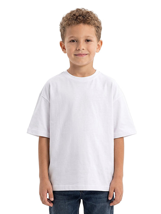 A young boy with curly light brown hair wearing an oversized white short-sleeve crewneck t-shirt and dark blue jeans, standing against a solid white studio background.