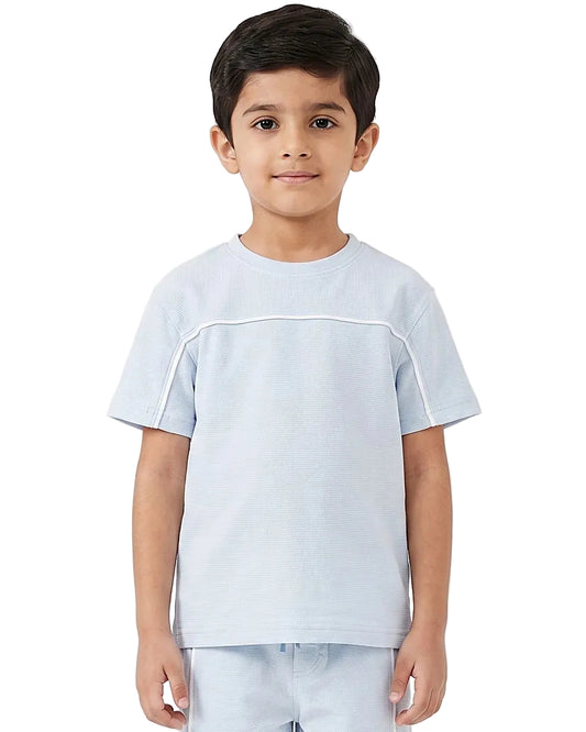 A young boy wearing a light blue short-sleeve t-shirt with white contrast stitching details across the chest and shoulders, paired with matching light blue pants, set against a plain white studio background.