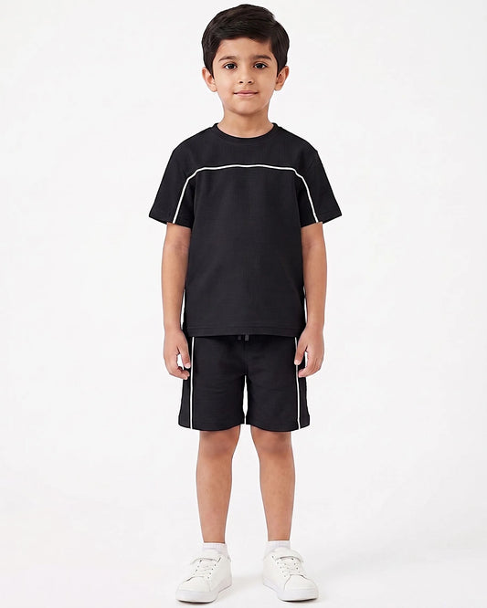 A young boy modeling a black short-sleeve t-shirt and matching black shorts with white contrast piping, paired with white sneakers, against a plain white studio background.