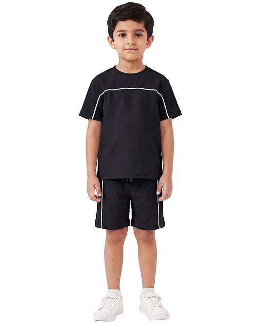 A young boy modeling a black short-sleeve t-shirt and matching black shorts with white contrast piping, paired with white sneakers, against a plain white studio background.