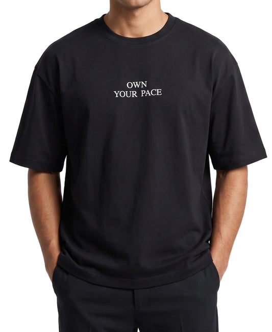 A person wearing an oversized black short-sleeve t-shirt with the text 'OWN YOUR PACE' printed in white serif letters on the chest, paired with black pants against a light gray background.