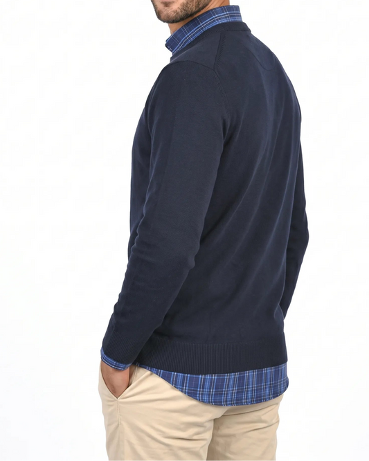 A man wearing a navy blue knit sweater over a blue and white plaid collared shirt, paired with beige trousers, photographed against a plain white background.