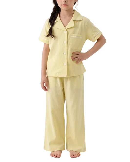 A young girl modeling a light yellow two-piece pajama set consisting of a short-sleeve button-up shirt and matching wide-leg pants, photographed against a seamless white studio background.