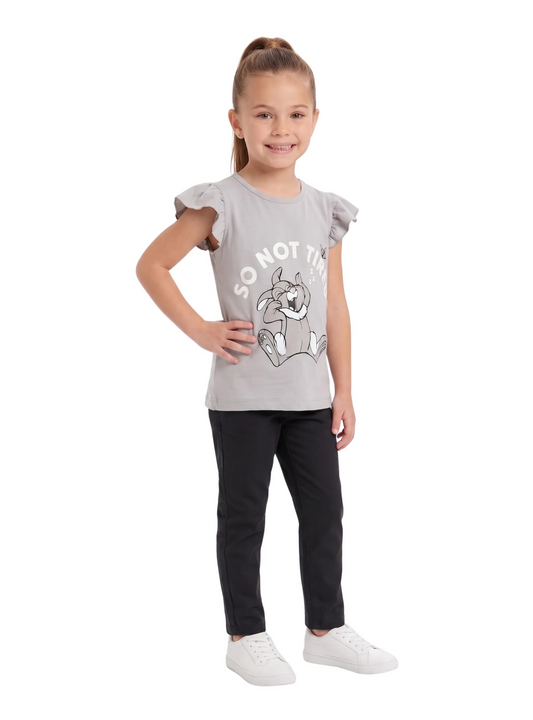 A young girl wearing a light grey t-shirt featuring a cartoon rabbit graphic and ruffle sleeves, paired with black pants and white sneakers