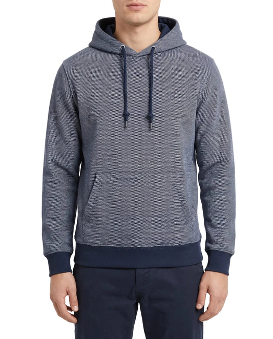A man wearing a textured blue-gray pullover hoodie with dark navy ribbed cuffs, hem, and drawstrings, paired with navy pants, against a plain light background.