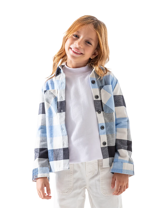 A young girl with light brown hair smiling, wearing a light blue, gray, and white plaid button-up shirt over a white turtleneck and white pants, photographed against a solid white background.