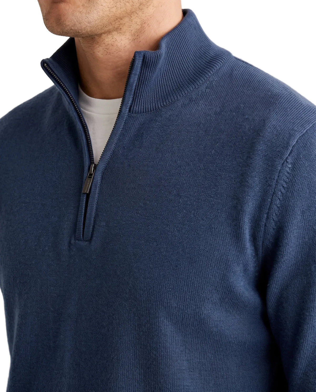 A man wearing a navy blue quarter-zip pullover sweater over a white undershirt and light beige trousers, standing against a plain white studio background.