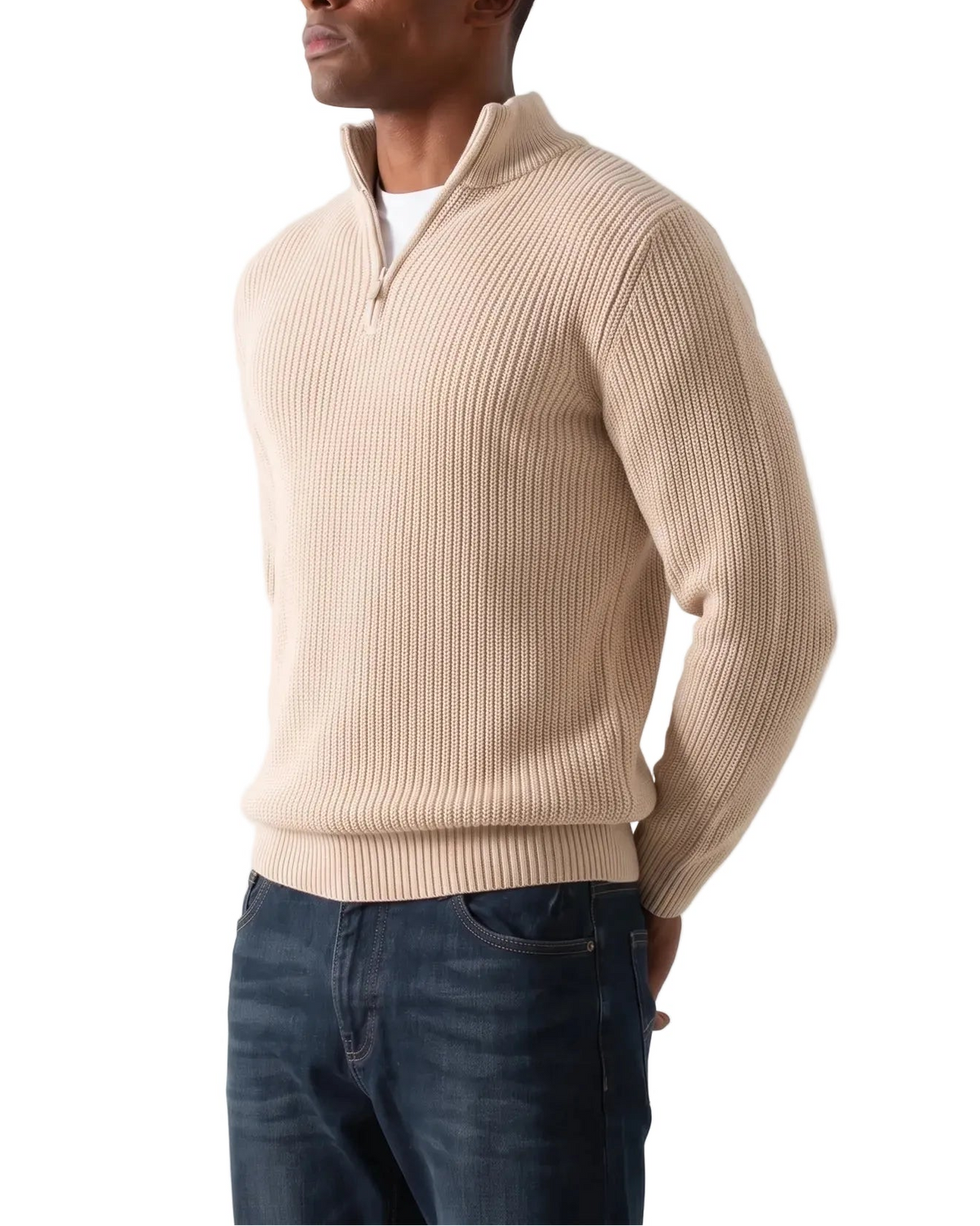 A man wearing a beige ribbed quarter-zip sweater over a white undershirt and dark blue jeans, standing against a plain light gray studio background.