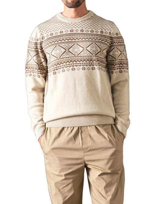 A man stands in front of a dark green velvet curtain wearing a beige crewneck sweater with a brown Nordic pattern, beige trousers, and white sneakers.