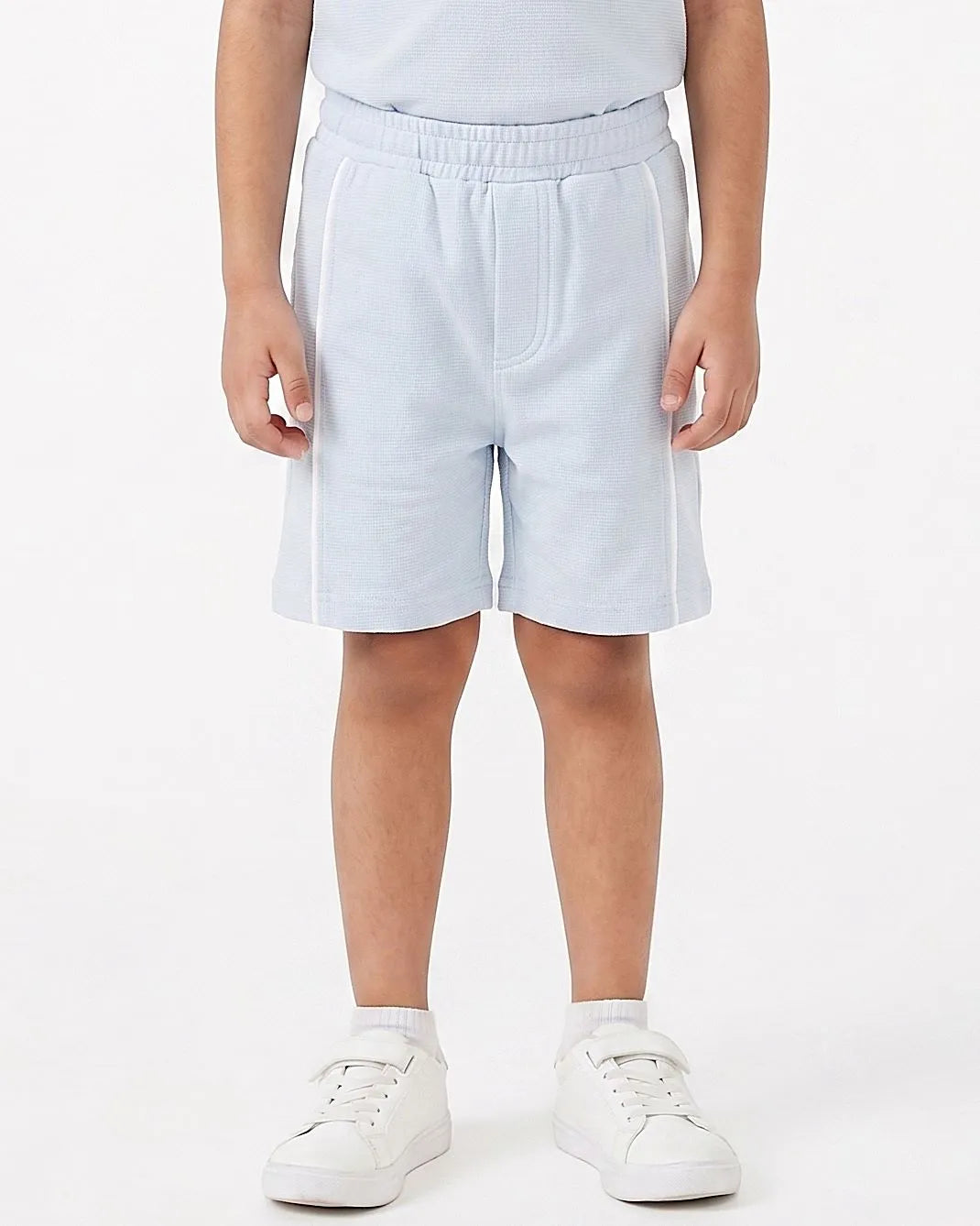 A child wearing light blue sweat shorts with white piping details on the sides, paired with white sneakers and a white top, shown from the waist down on a white studio background.