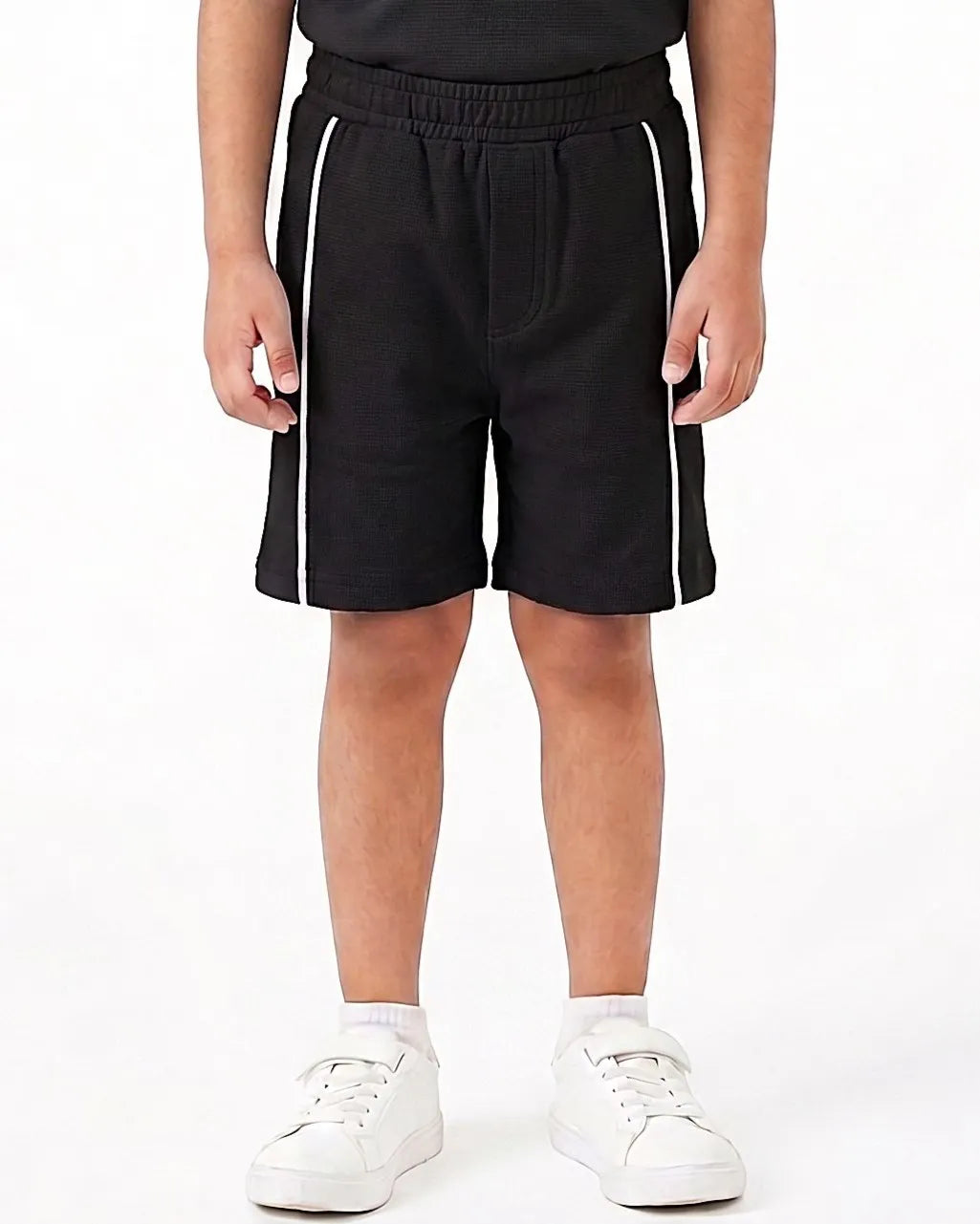 A child wearing black athletic shorts with white side stripes, paired with a black top and white sneakers, shown from the waist down on a white studio background.