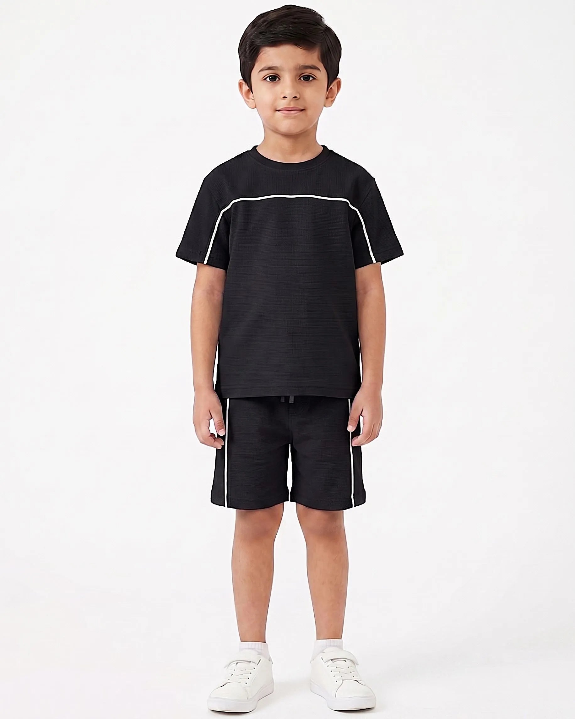 A young boy modeling a black short-sleeve t-shirt and matching black shorts with white contrast piping, paired with white sneakers, against a plain white studio background.