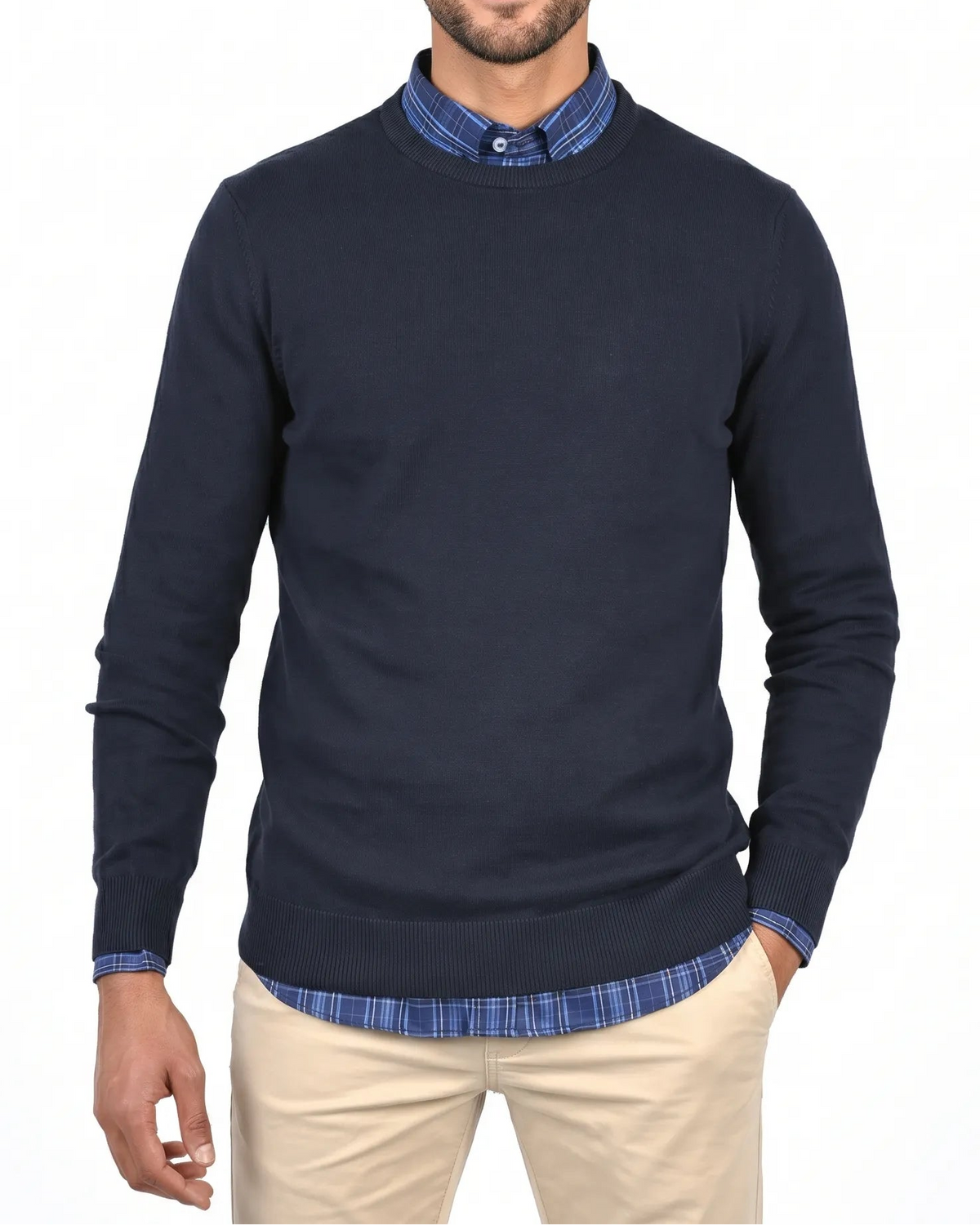 A man wearing a navy blue long-sleeve knit sweater over a blue and white checkered collared shirt, paired with beige trousers, against a plain white background.