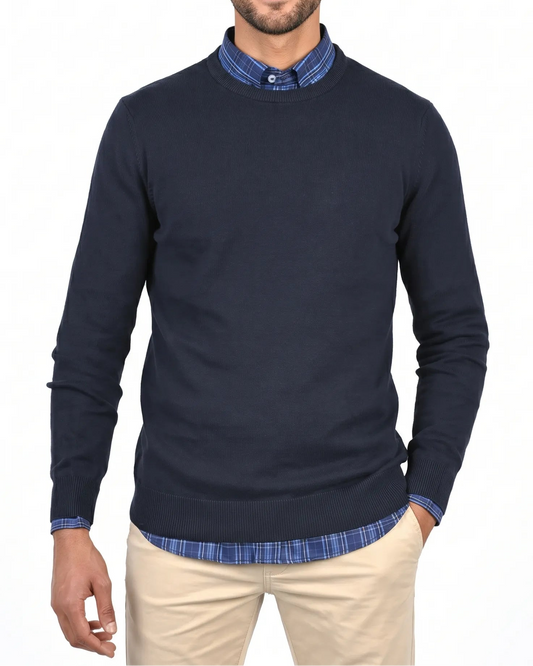 A man wearing a navy blue long-sleeve knit sweater over a blue and white checkered collared shirt, paired with beige trousers, against a plain white background.