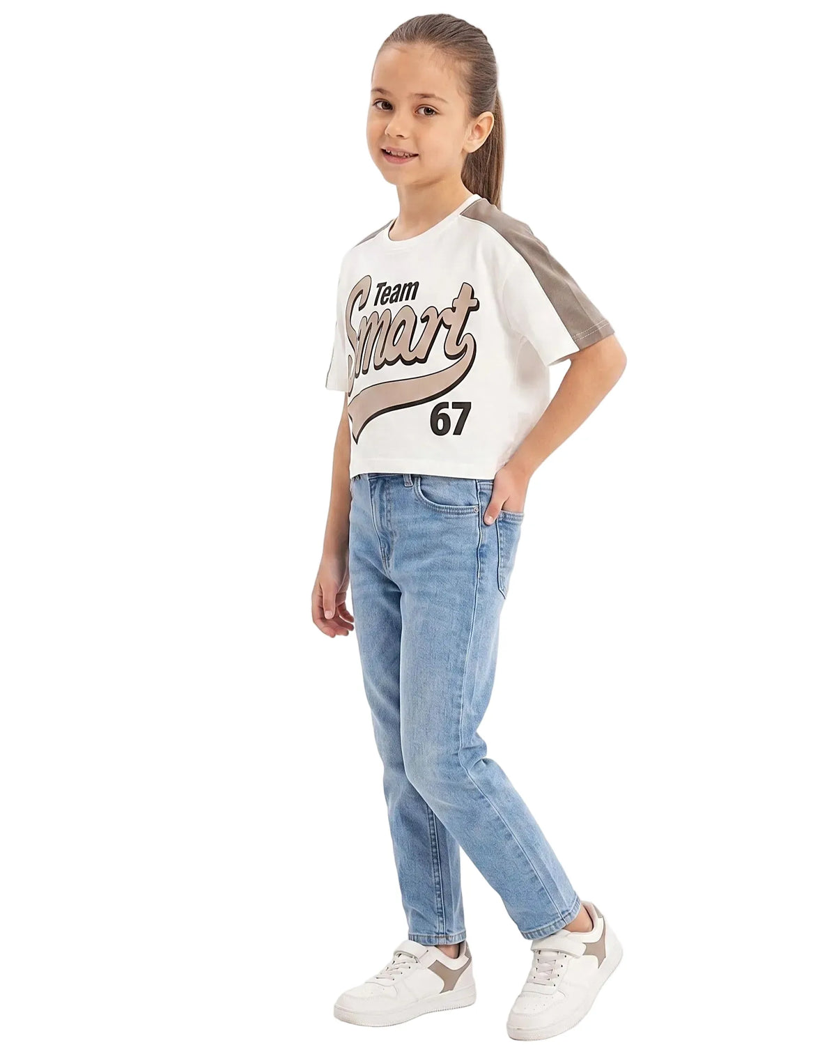 Full-body side view of a young girl modeling a Girls' "Team Smart 67" Print T-Shirt with contrast shoulders, paired with blue jeans and white sneakers.