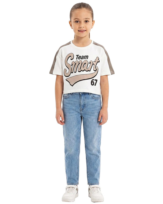 Young girl modeling a Girls' 'Team Smart 67' Print Cotton Blend T-Shirt in off-white with contrast shoulders, paired with blue jeans and white sneakers.