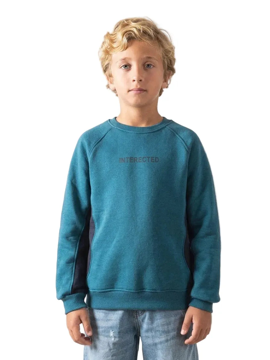 A young boy with curly blonde hair wearing a teal blue crewneck sweatshirt with 'INTERACTED.' printed in dark gray on the chest, paired with light blue jeans, standing against a plain white background.