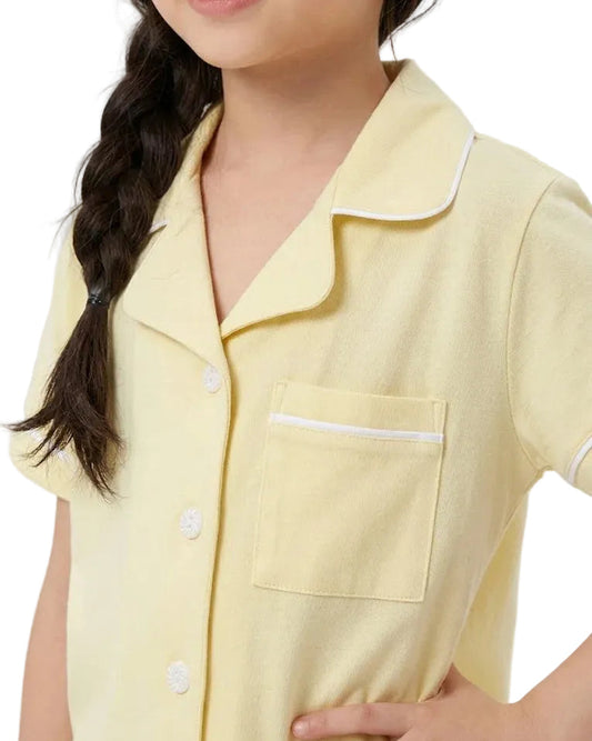 A close-up of a child wearing a pale yellow short-sleeve pajama top with white piping and a chest pocket, photographed against a plain light background.