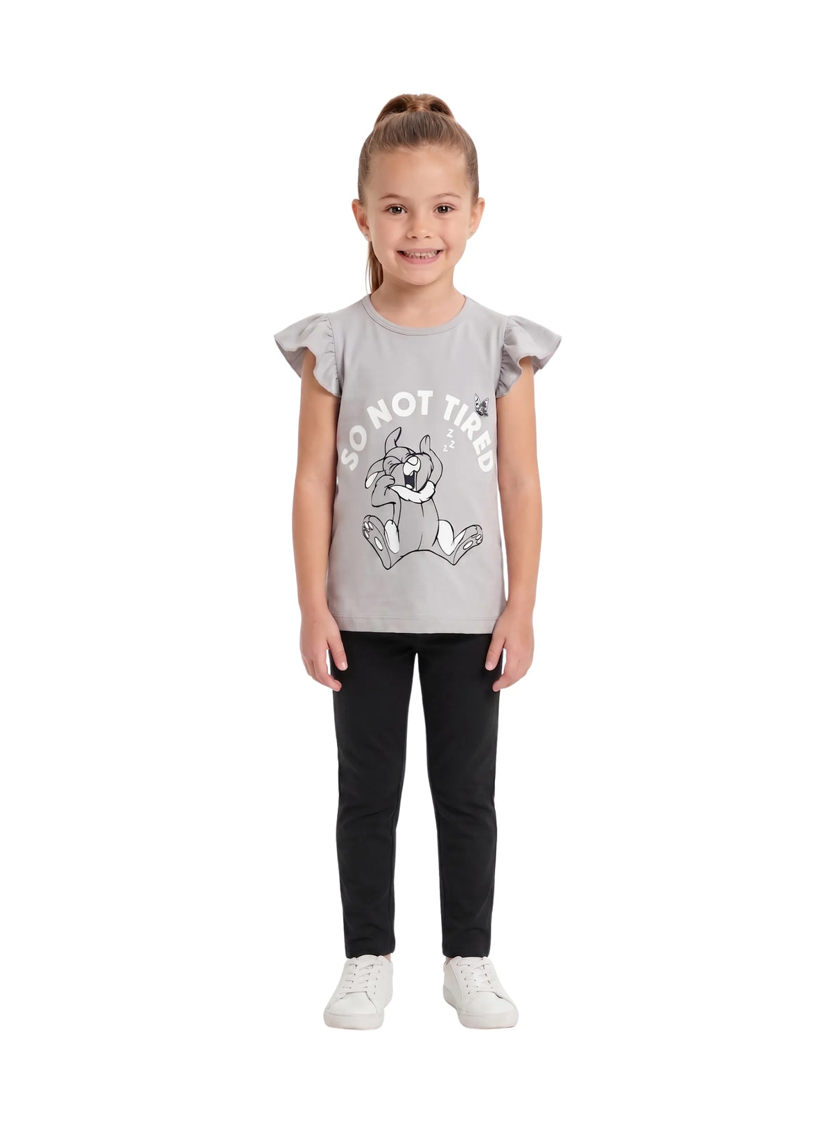A young girl wearing a light grey t-shirt featuring a cartoon rabbit graphic and ruffle sleeves, paired with black pants and white sneakers