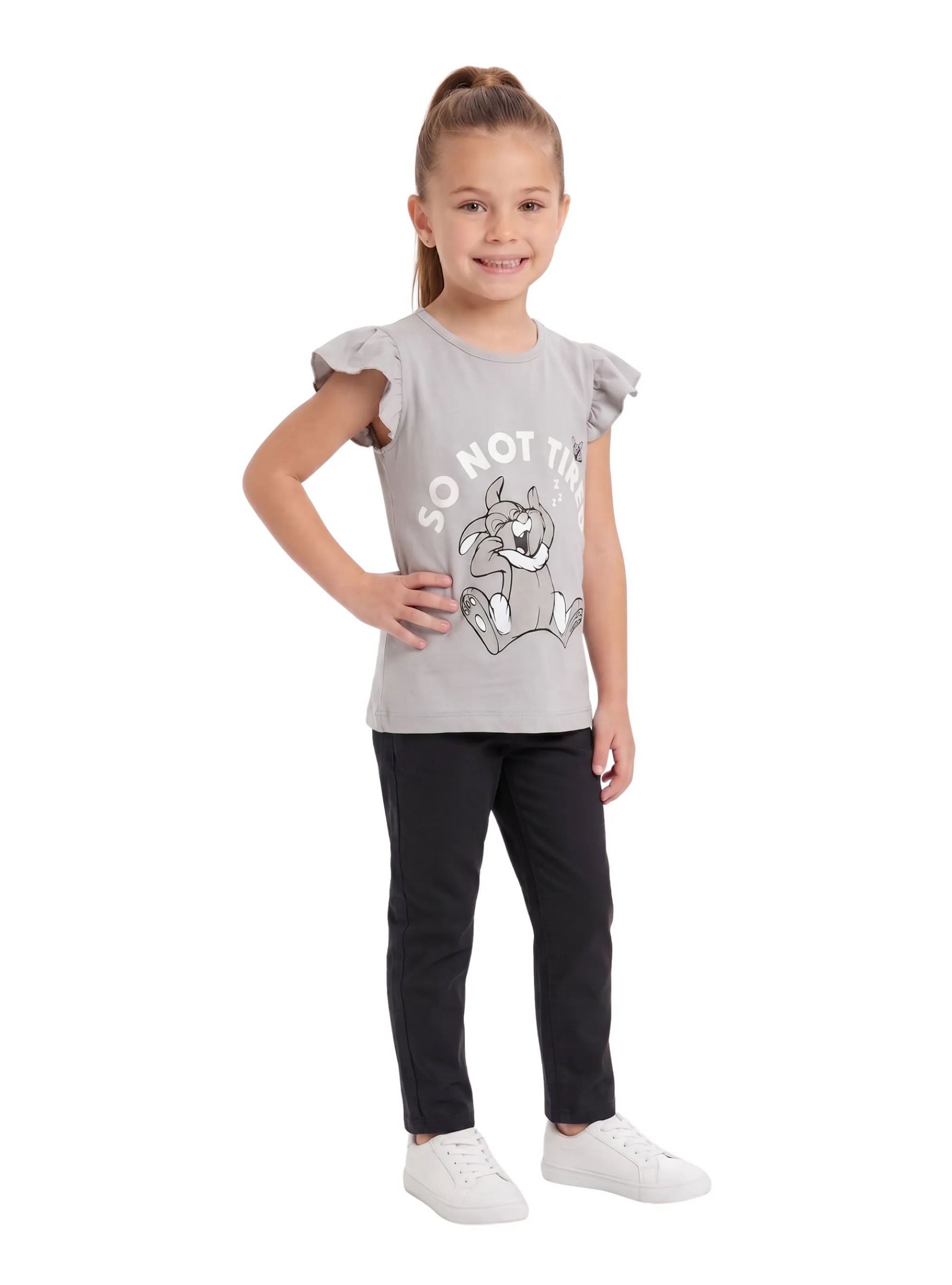 A young girl wearing a light grey t-shirt featuring a cartoon rabbit graphic and ruffle sleeves, paired with black pants and white sneakers