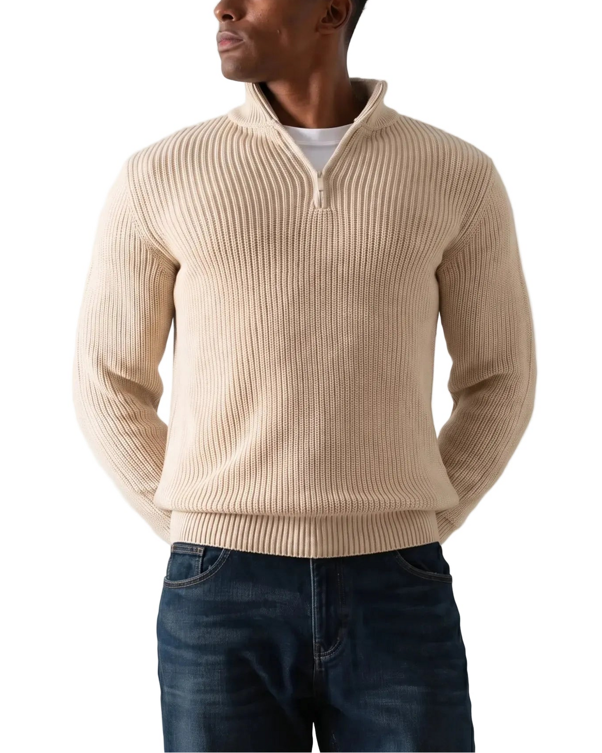 A man wearing a beige ribbed quarter-zip sweater over a white undershirt and dark blue jeans, standing against a plain light gray background.