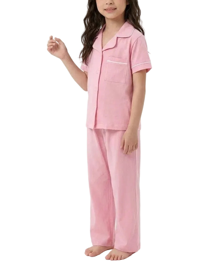 A young girl modeling a two-piece pink pajama set consisting of a short-sleeve button-up top with a chest pocket and matching wide-leg pants, photographed against a plain white studio background.