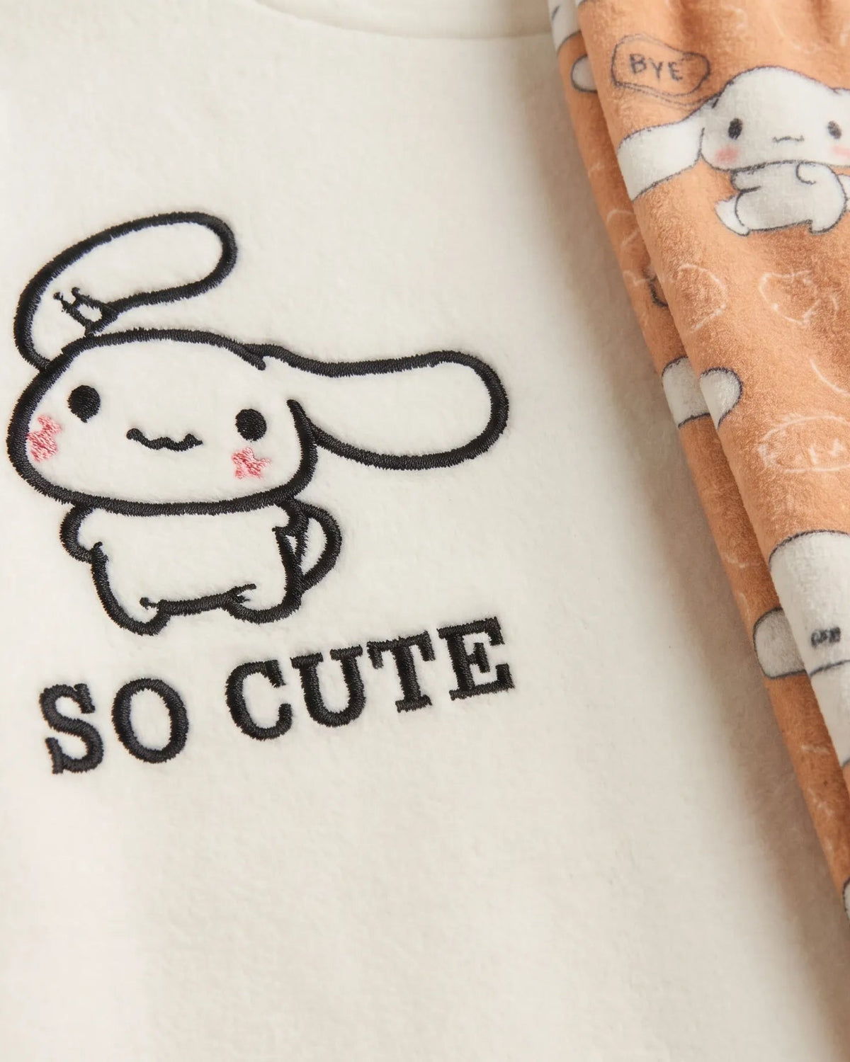 Close-up of a soft fabric garment showing a cream-colored section with a black cartoon bunny outline and the words 'SO CUTE', next to peach-colored fabric with a white bunny print.