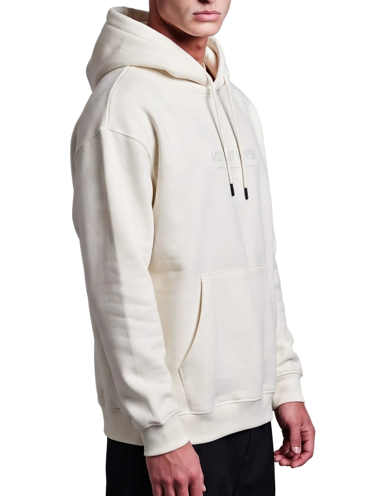 A man wearing a cream-colored hoodie with a tonal chest logo, shown from a side profile against a neutral gray background.