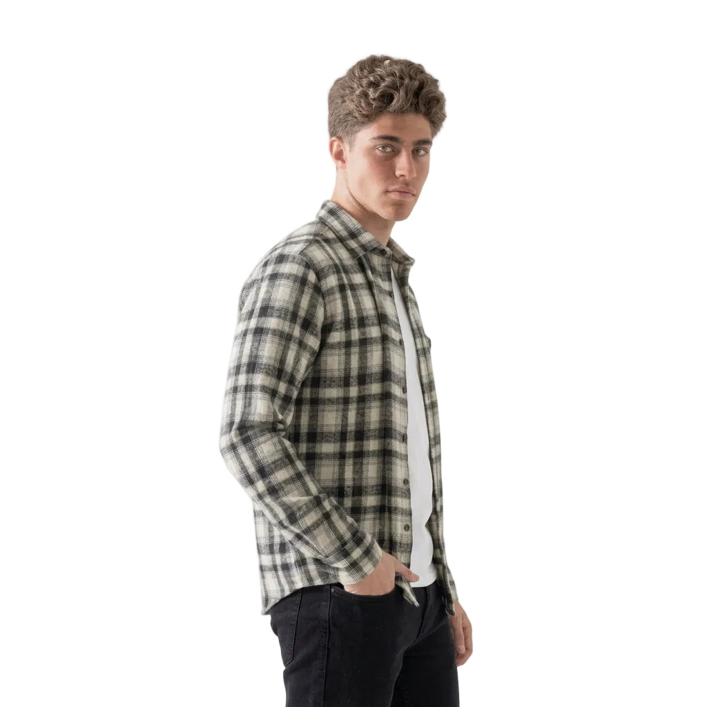 A young man with curly brown hair is shown in profile wearing a black and beige plaid button-up shirt over a white t-shirt and dark jeans, photographed against a solid white background.