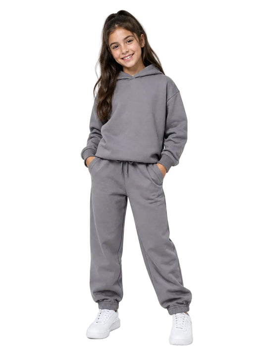 A young girl modeling a matching gray two-piece loungewear set, consisting of a pullover hoodie and jogger sweatpants, paired with white sneakers against a plain light gray studio background.