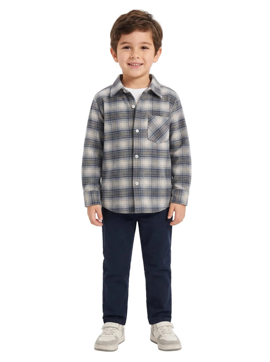 A young boy stands against a light gray studio background wearing a long-sleeve plaid button-up shirt in gray, navy, and white, paired with dark navy pants and light gray sneakers.