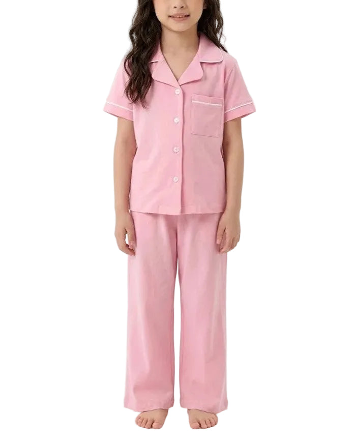 A young girl modeling a two-piece pink pajama set consisting of a short-sleeve button-up top with white piping and matching wide-leg pants, shot against a plain white studio background.