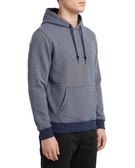 A man wearing a textured heathered blue hoodie with dark navy ribbed cuffs, hem, and drawstrings, paired with black pants, photographed against a clean white studio background.