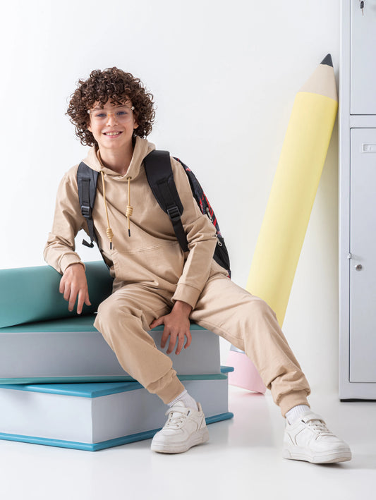 Person in beige hoodie and pants with a backpack standing next to books and lockers.