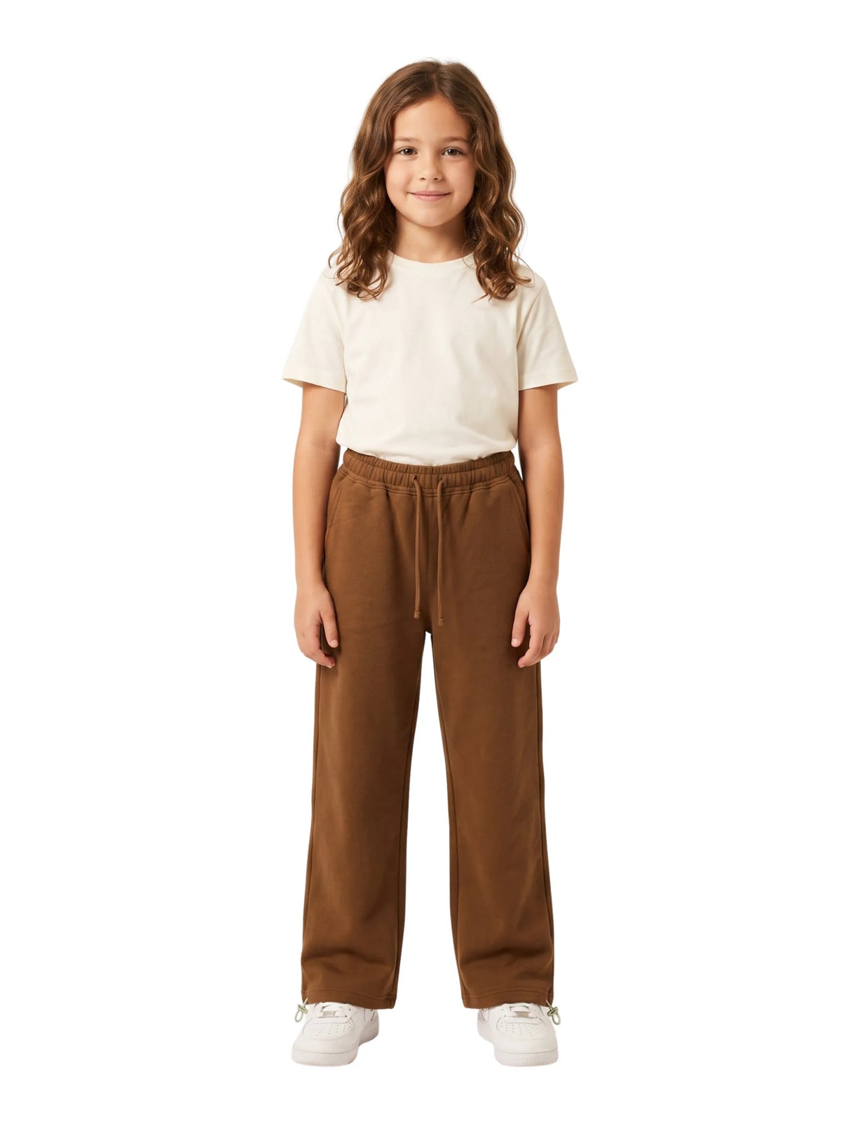 A young girl with wavy brown hair stands against a solid white background, wearing a cream short-sleeve t-shirt, brown wide-leg drawstring pants, and white sneakers.